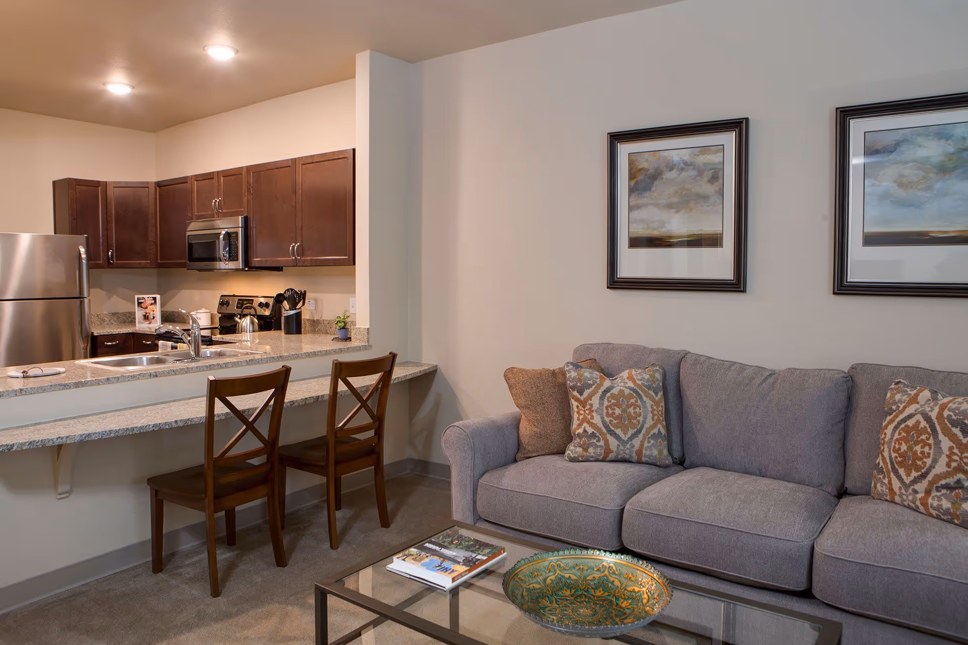 A cozy living area with a gray sofa adorned with patterned cushions, a glass coffee table with a decorative bowl and a book, and a kitchen in the background featuring wooden cabinets, a stainless steel refrigerator, microwave, stove, and a countertop with two wooden chairs.