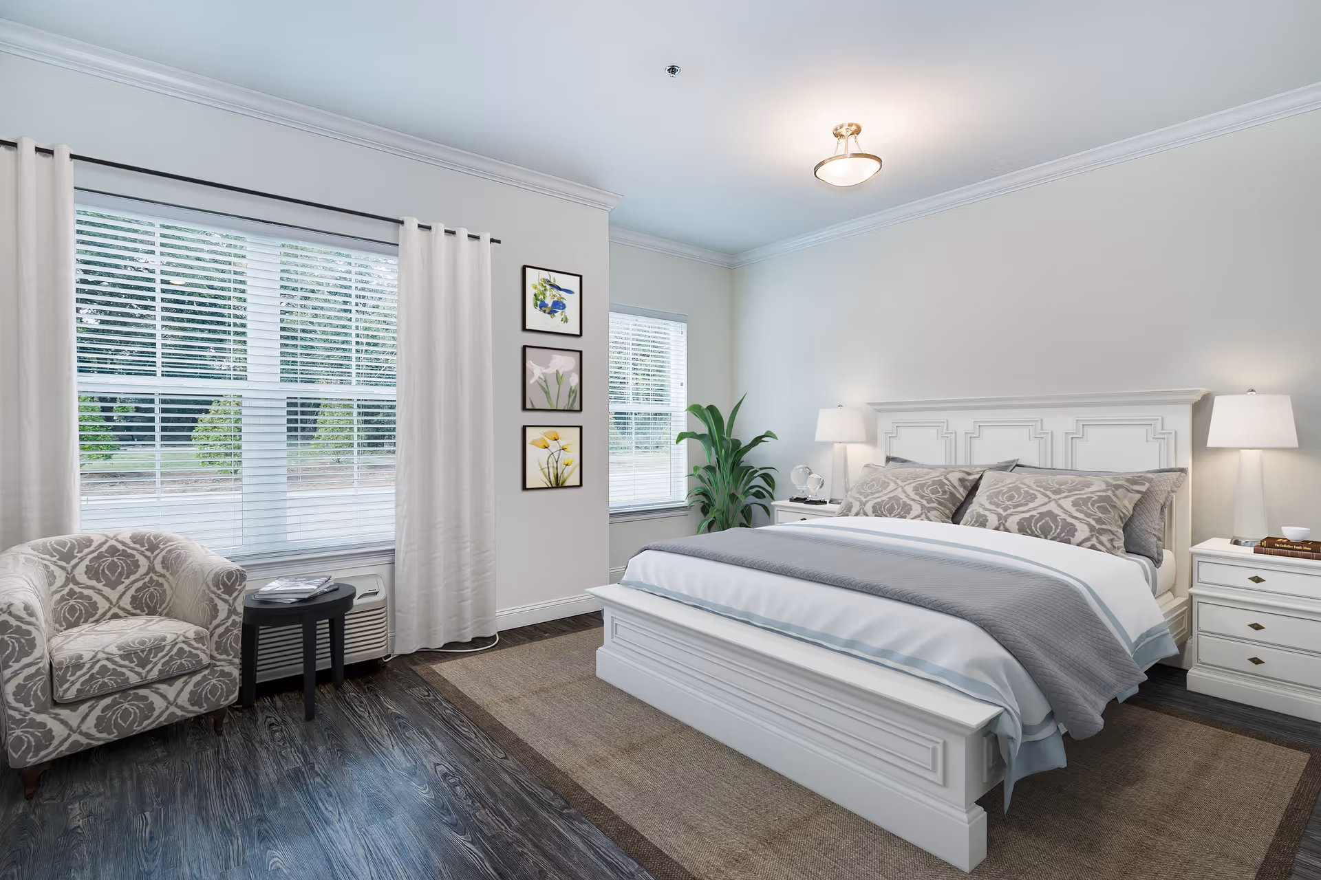 A bright and spacious bedroom with a large white bed featuring patterned pillows and a gray blanket. The room has two white nightstands with lamps on each side of the bed, a patterned armchair next to a small round black table, large windows with white blinds and curtains, three framed floral artworks on the wall, and a green potted plant in the corner. The floor is dark wood with a large beige rug under the bed.