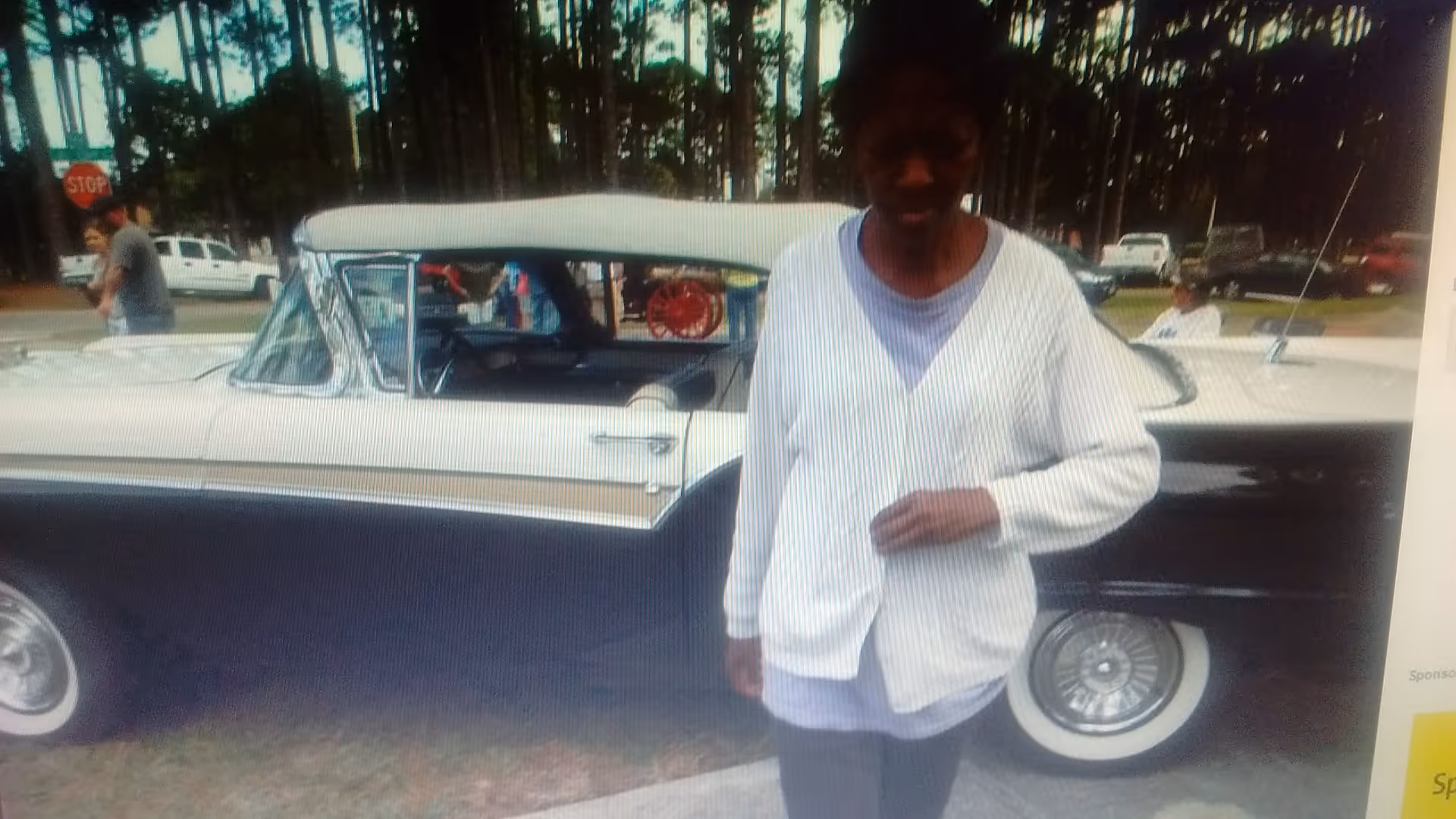 A person wearing a white cardigan and gray pants standing next to a vintage black and white car parked outdoors with trees and other vehicles in the background.
