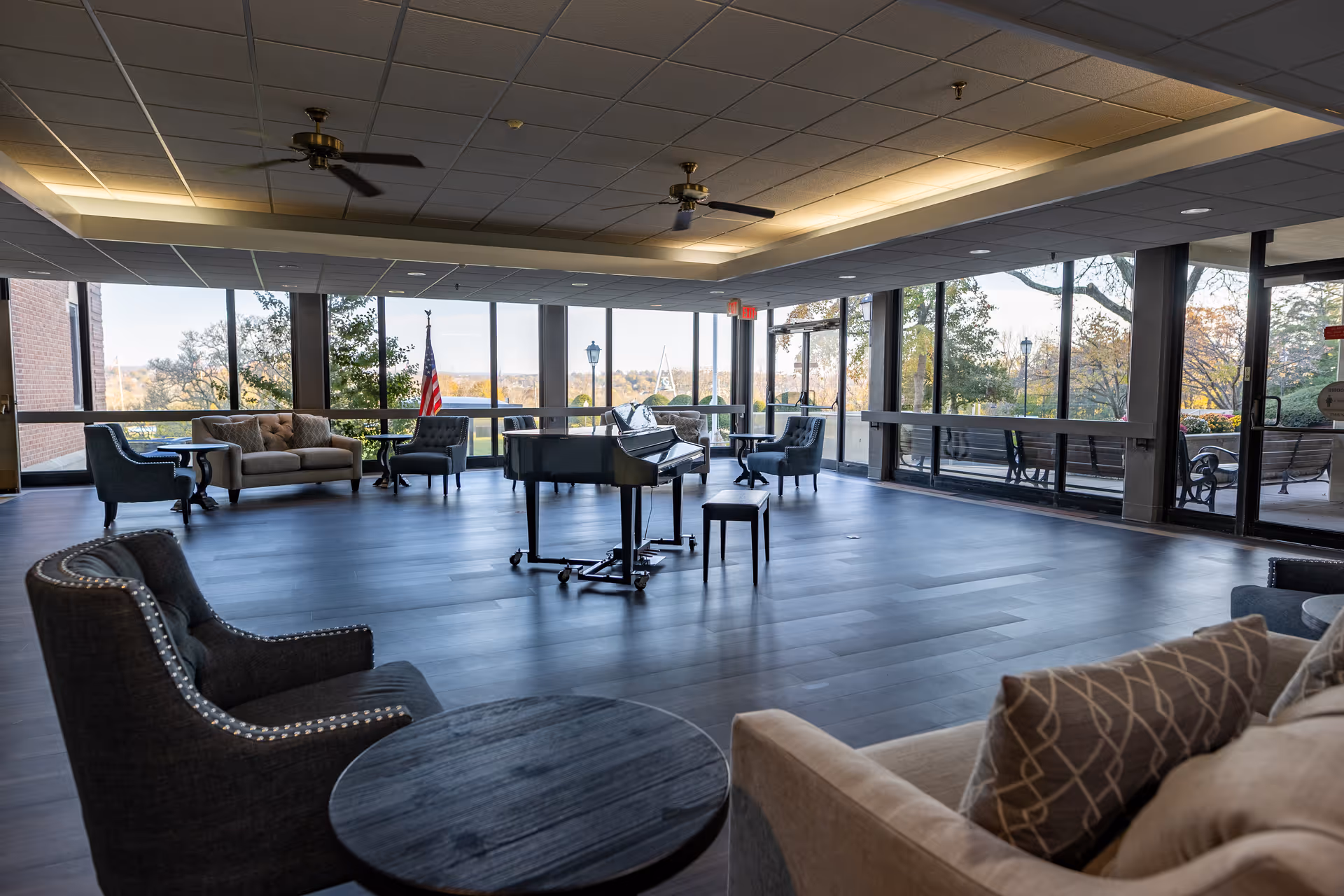 A spacious lounge area with large windows offering a view of trees and outdoor benches. The room features a grand piano in the center, several upholstered chairs and sofas arranged around small round tables, ceiling fans, and soft overhead lighting.