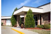 Brick single-story healthcare facility entrance with a covered portico, evergreen shrubs, and a curved driveway under a blue sky.