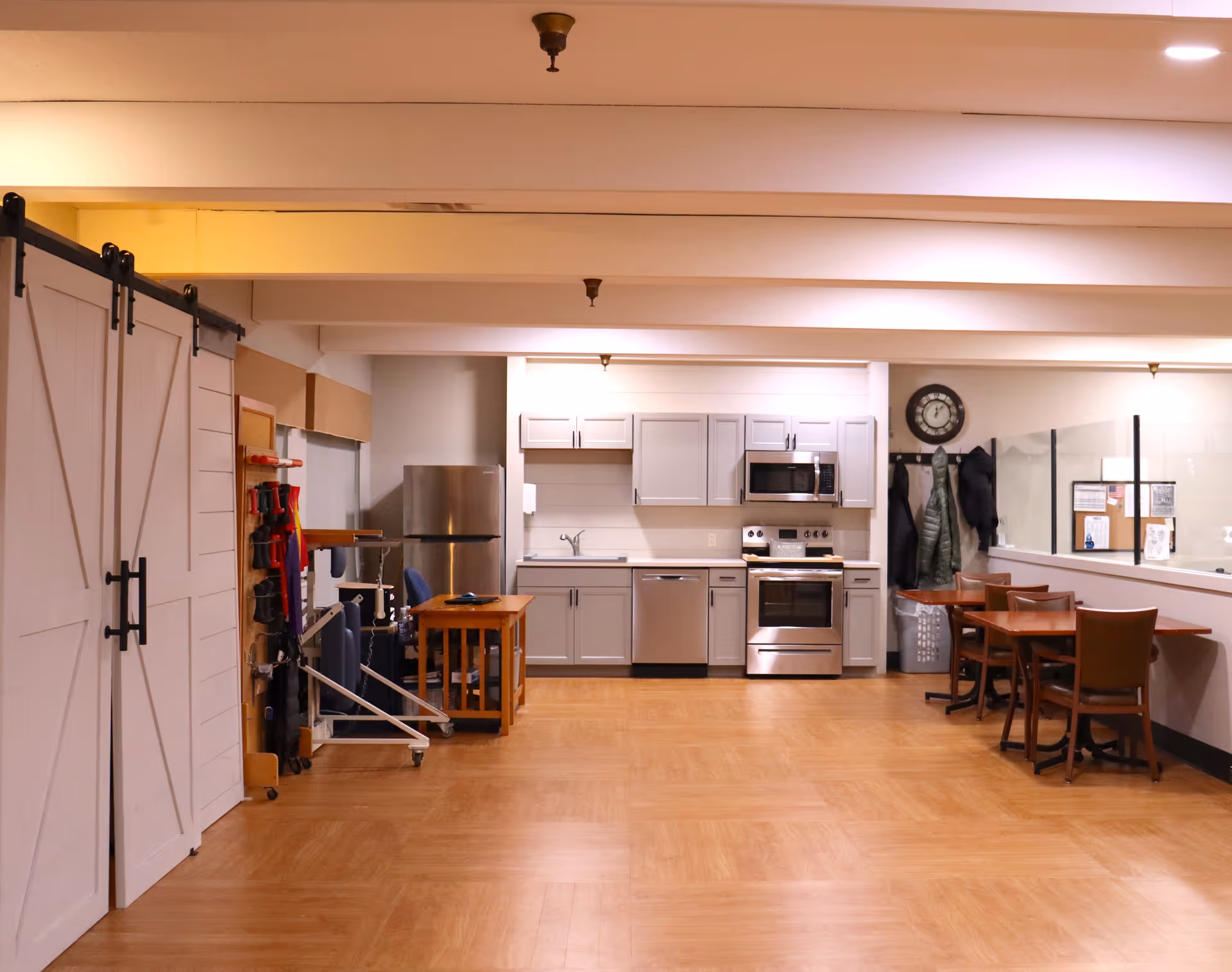 Interior view of a senior living facility kitchen and dining area with light wood flooring, white cabinets, stainless steel appliances including a refrigerator, stove, microwave, and dishwasher. There are two small wooden tables with chairs on the right side, a coat rack with jackets and a clock above it, and sliding barn doors on the left side.