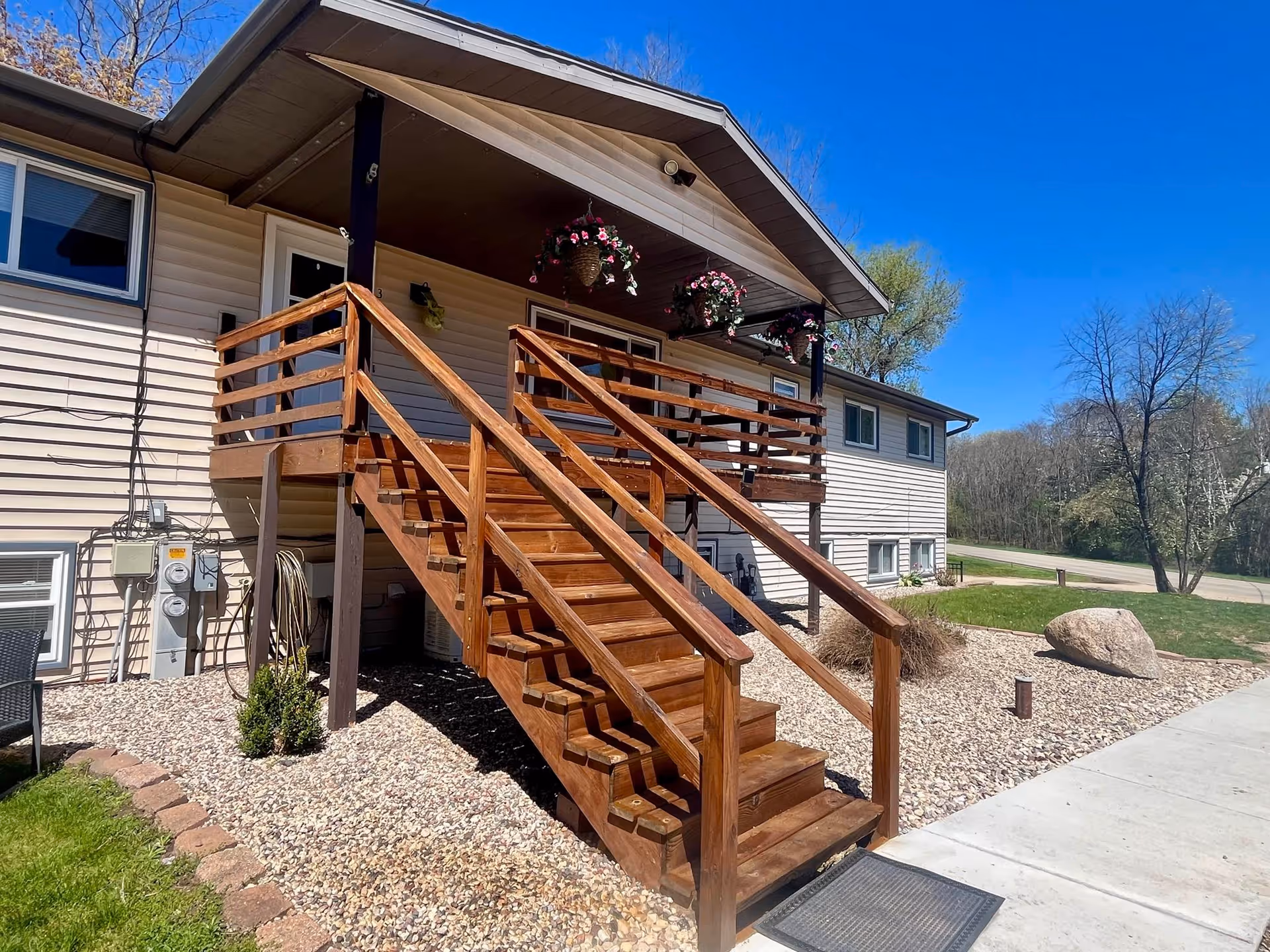 Exterior view of a beige two-story building with a wooden staircase leading to a covered porch decorated with hanging flower baskets. The area around the building is landscaped with rocks, a large boulder, and some greenery under a clear blue sky.