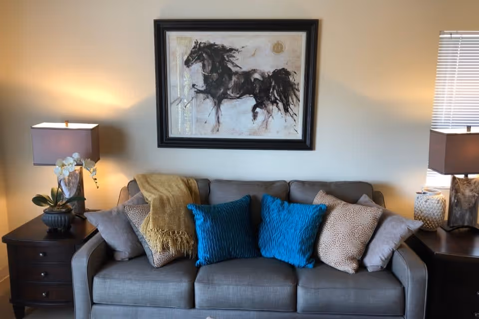A cozy living room area with a gray sofa adorned with multiple cushions in blue, beige, and gray colors, and a mustard yellow throw blanket. On either side of the sofa are dark wooden side tables with table lamps and decorative items. Above the sofa hangs a framed painting of a black horse.