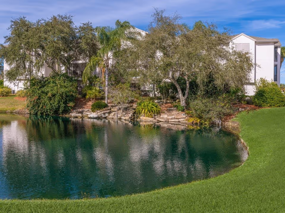 A pond and landscaped lawn in front of a multi-story white residential building partially obscured by trees.