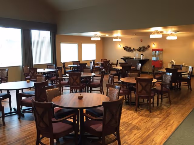 A dining room with multiple round wooden tables and chairs arranged neatly. The room has large windows with curtains allowing natural light to enter. In the background, there is a counter with a coffee machine and a red popcorn machine. The floor is wooden and the walls are painted beige.