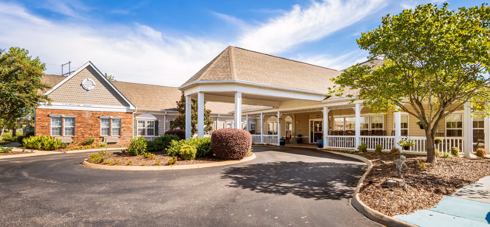 Exterior view of a senior living facility named The Neighborhood at Madison, showing a covered entrance with white pillars, a circular driveway, landscaped bushes, trees, and a clear sky.