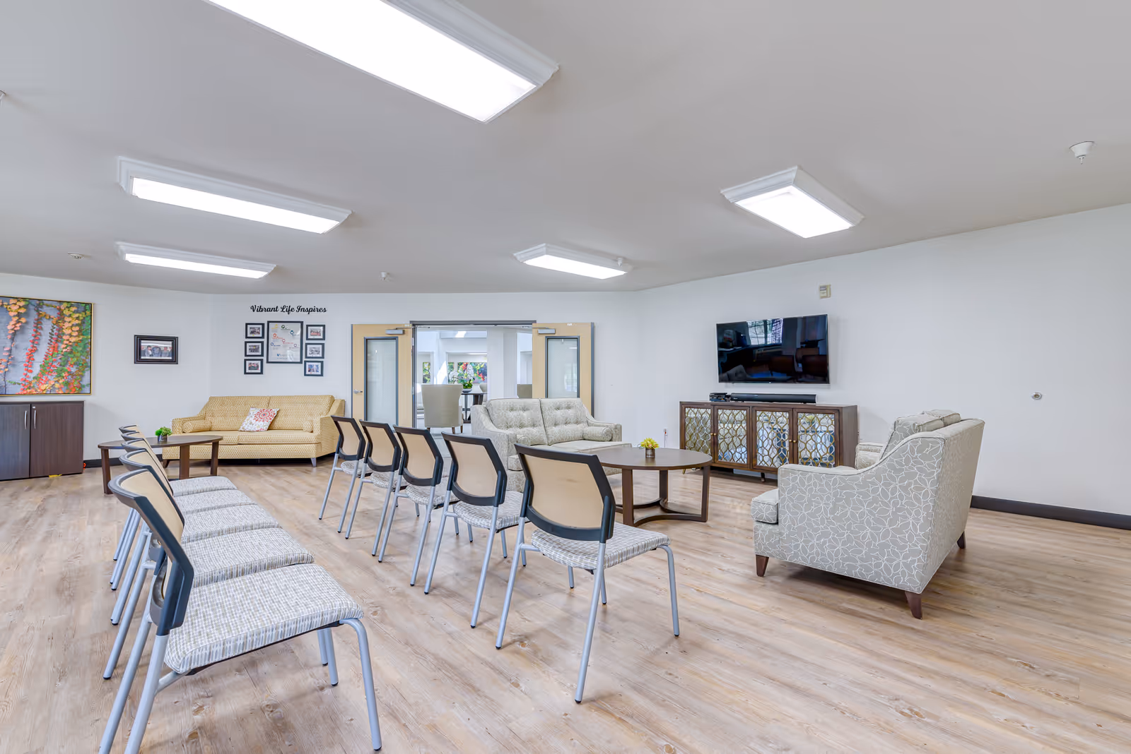 A spacious senior living common area with multiple chairs arranged in rows facing a seating area with two sofas, an armchair, and a round coffee table. A flat-screen TV is mounted on the wall above a decorative cabinet. The room has wood flooring, white walls, and bright overhead lighting. There is artwork and framed photos on the walls, and double doors lead to another room.