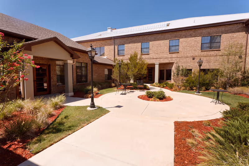A sunny landscaped courtyard with paved walkways, outdoor seating, lamp posts, and a two-story brick building.