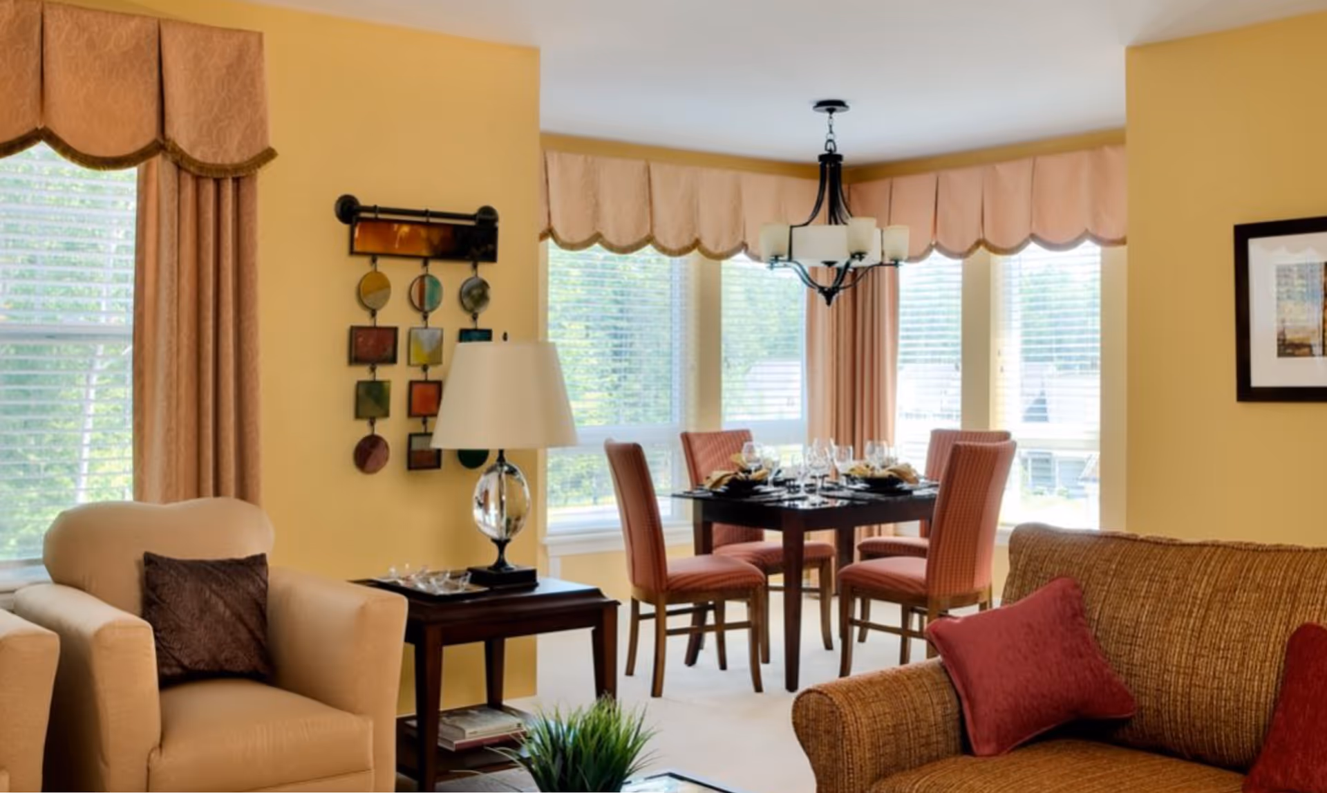 A cozy living and dining area with beige walls and large windows covered by peach-colored valances and curtains. The living area has a beige armchair with a dark brown pillow, a brown sofa with red pillows, and a dark wooden side table with a lamp and decorative items. The dining area features a dark wooden table set with plates, glasses, and napkins, surrounded by four upholstered chairs in a reddish pattern. A black chandelier hangs above the dining table.