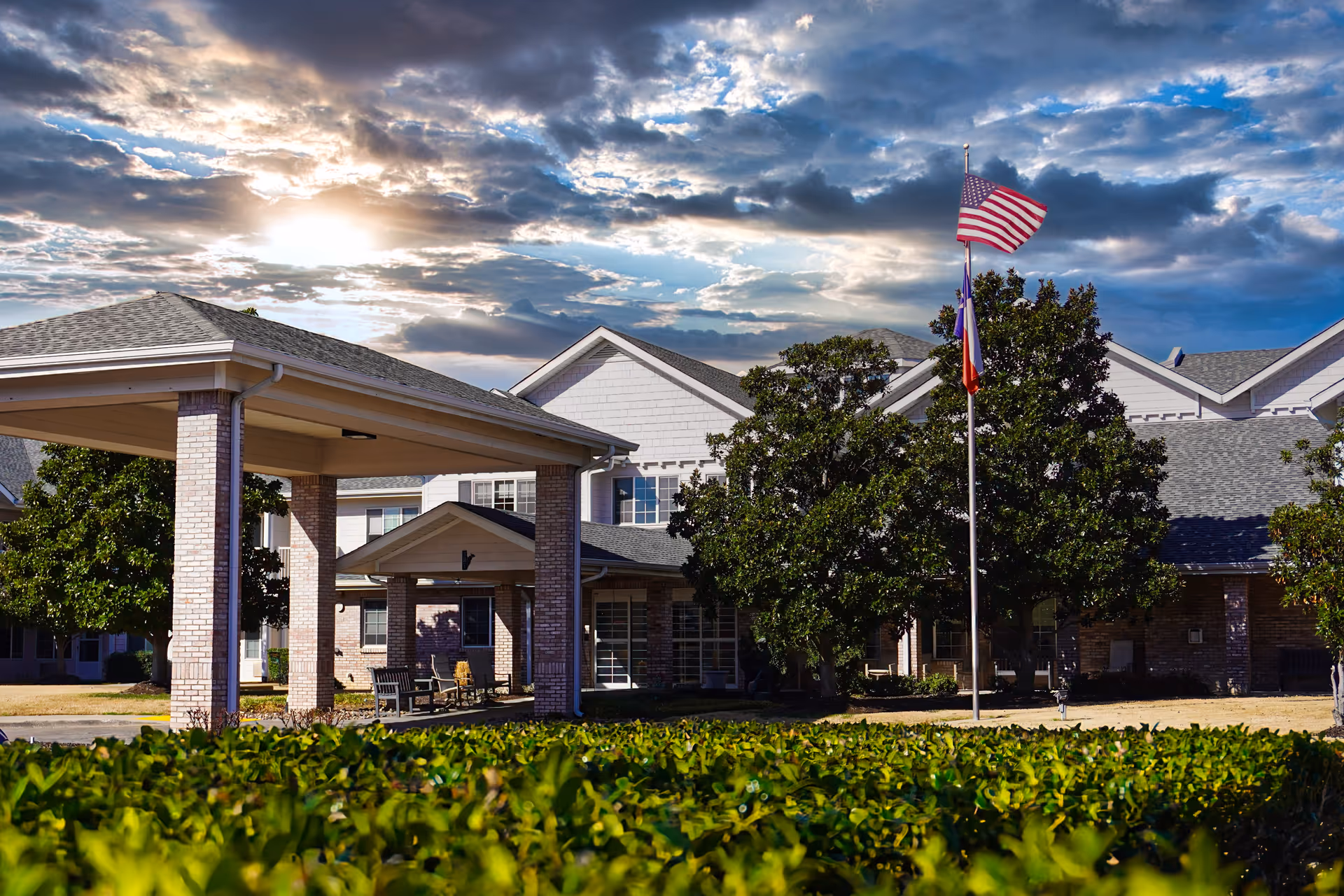 Exterior view of Pinewood Hills - Sky Active Living facility with a covered entrance, trees, and an American flag on a flagpole under a partly cloudy sky during sunset.