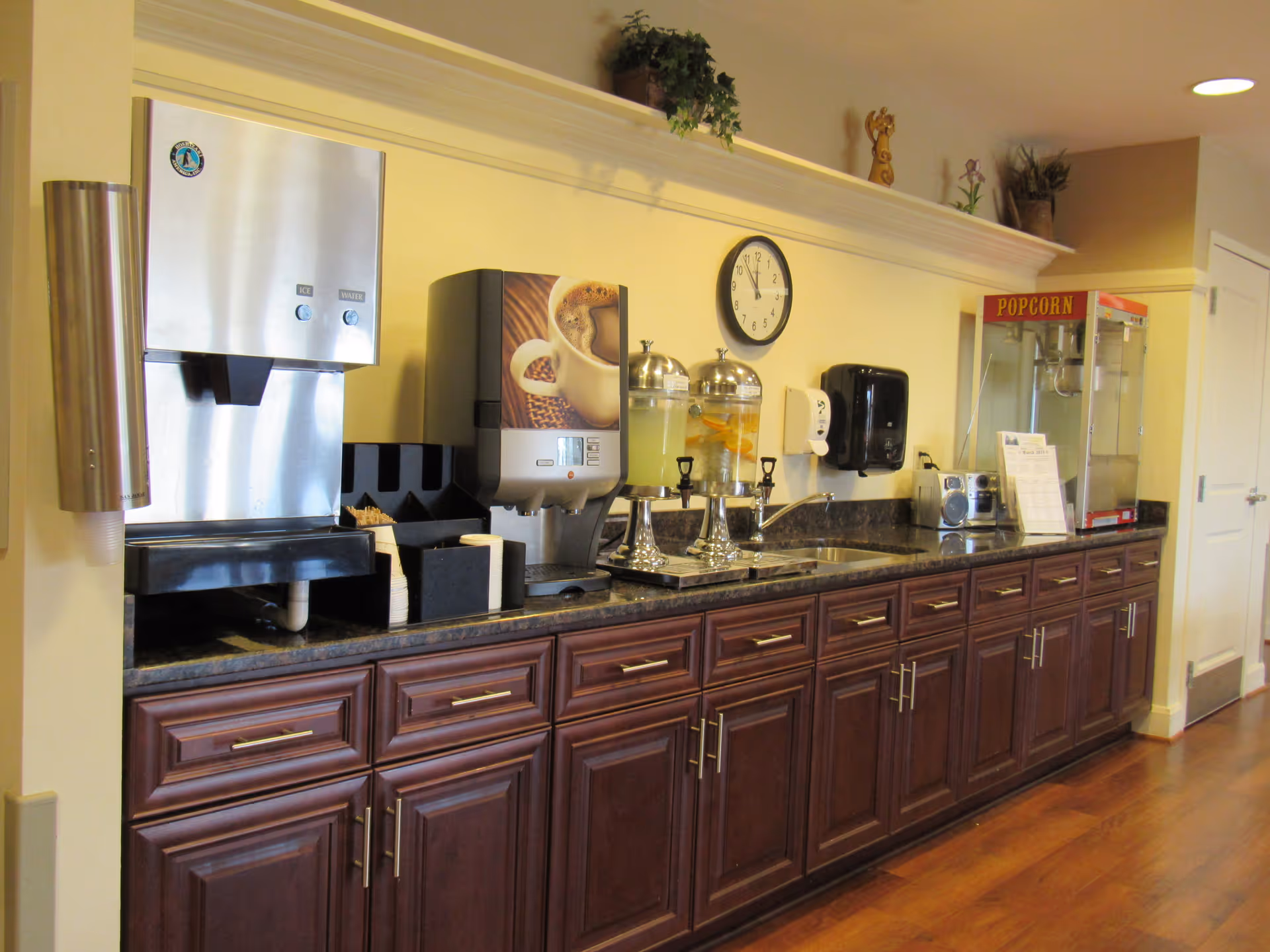 A beverage and snack station with a stainless steel water dispenser, a coffee machine, two glass beverage dispensers with lemonade and infused water, a sink, a popcorn machine, and wooden cabinets below. A clock is mounted on the wall above the counter, and there are decorative plants on a shelf above.