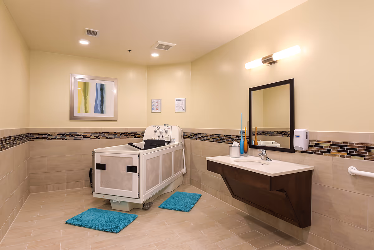 A spacious, well-lit bathroom with beige tiled walls and floor. The room features a large, specialized bathing tub with safety handles, two blue bath mats on the floor, a wall-mounted sink with a dark wood cabinet, a rectangular mirror above the sink, a soap dispenser, and modern light fixtures. There is also a colorful abstract painting on the wall.
