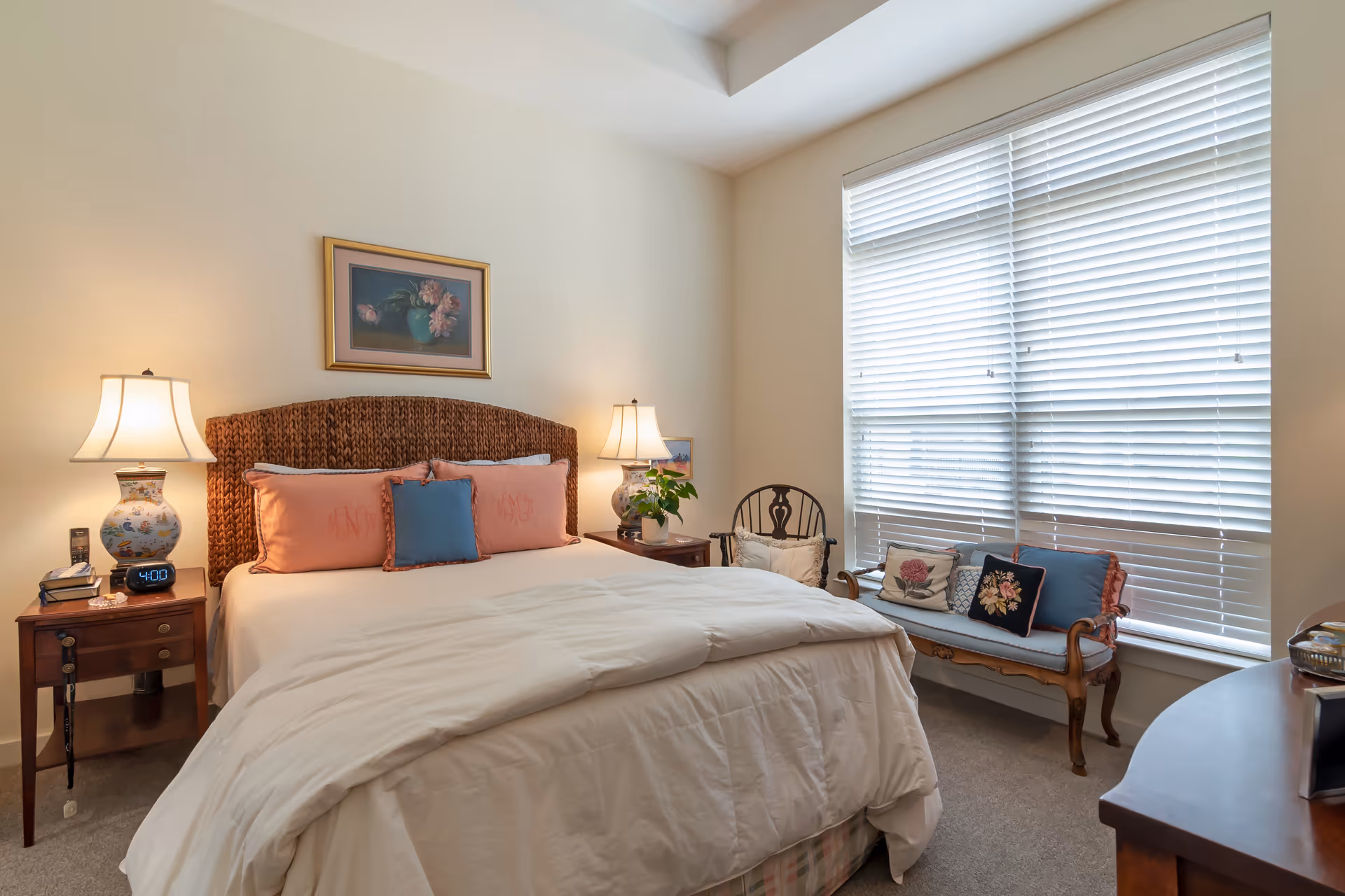 A cozy bedroom with a large bed featuring a woven headboard, white bedding, and decorative pillows. There are two bedside tables with matching lamps, a digital alarm clock, and a small plant. A wooden chair with a cushion and a wooden bench with several decorative pillows are placed near a large window with white blinds. A framed floral painting hangs above the bed.