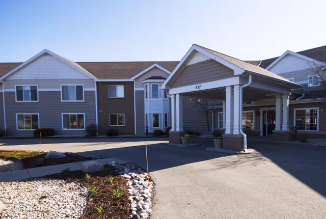 Exterior view of a senior living facility building with a covered entrance and driveway. The building has two stories with multiple windows, beige and white siding, and a landscaped area with rocks and plants in the foreground.