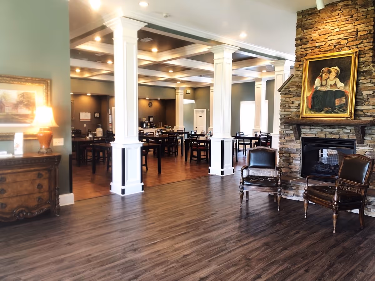 Spacious common room with wood floors, white columns, dining tables, and a stone fireplace with chairs and framed artwork.