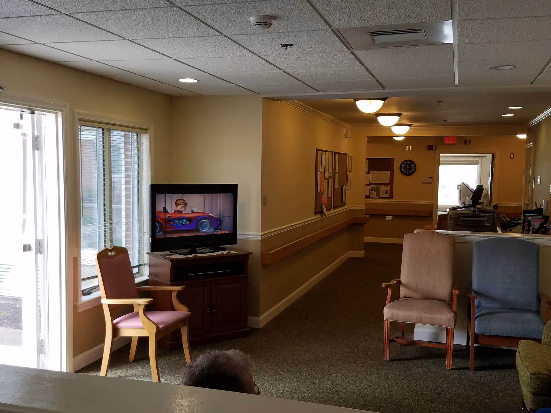 Interior view of a senior living facility hallway with several chairs arranged near a television playing a cartoon. The hallway has bulletin boards on the wall, ceiling lights, and a window letting in natural light.