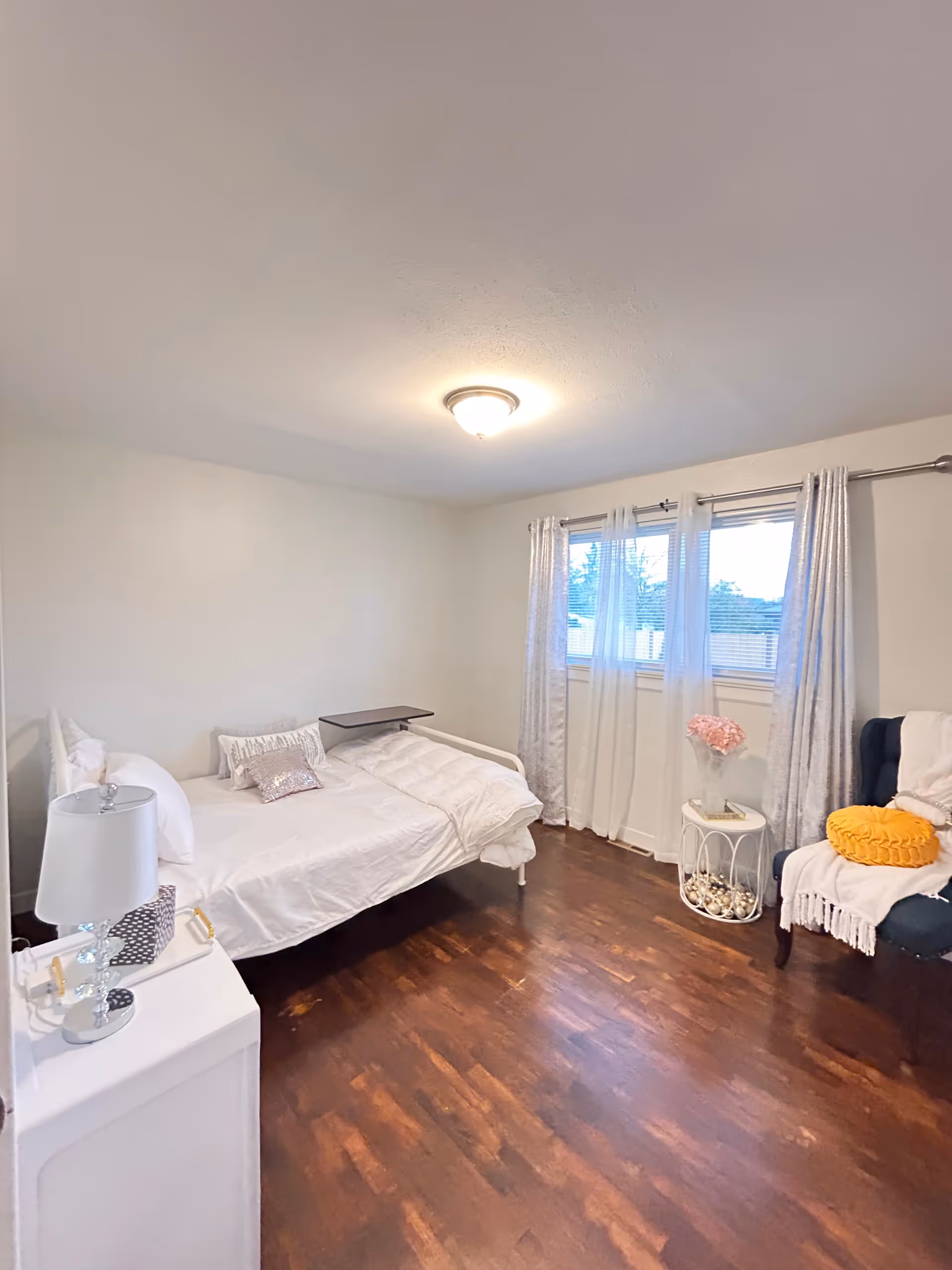 A bright bedroom with a single white bed covered with white bedding and decorative pillows. Next to the bed is a white nightstand with a lamp and some items on it. A window with sheer white curtains lets in natural light. A small round table with a vase of pink flowers and decorative items is beside a dark upholstered chair with a yellow round pillow and a white throw blanket. The room has wooden flooring and a ceiling light fixture.