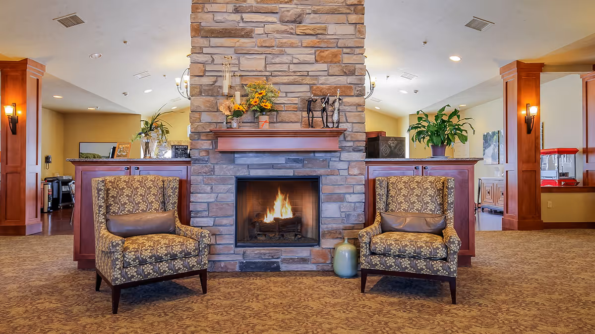 A cozy interior lounge area with a stone fireplace in the center, flanked by two patterned armchairs with brown cushions. The fireplace mantel is decorated with flowers, candles, and small sculptures. The room has warm lighting with wooden columns and plants on either side behind the chairs.