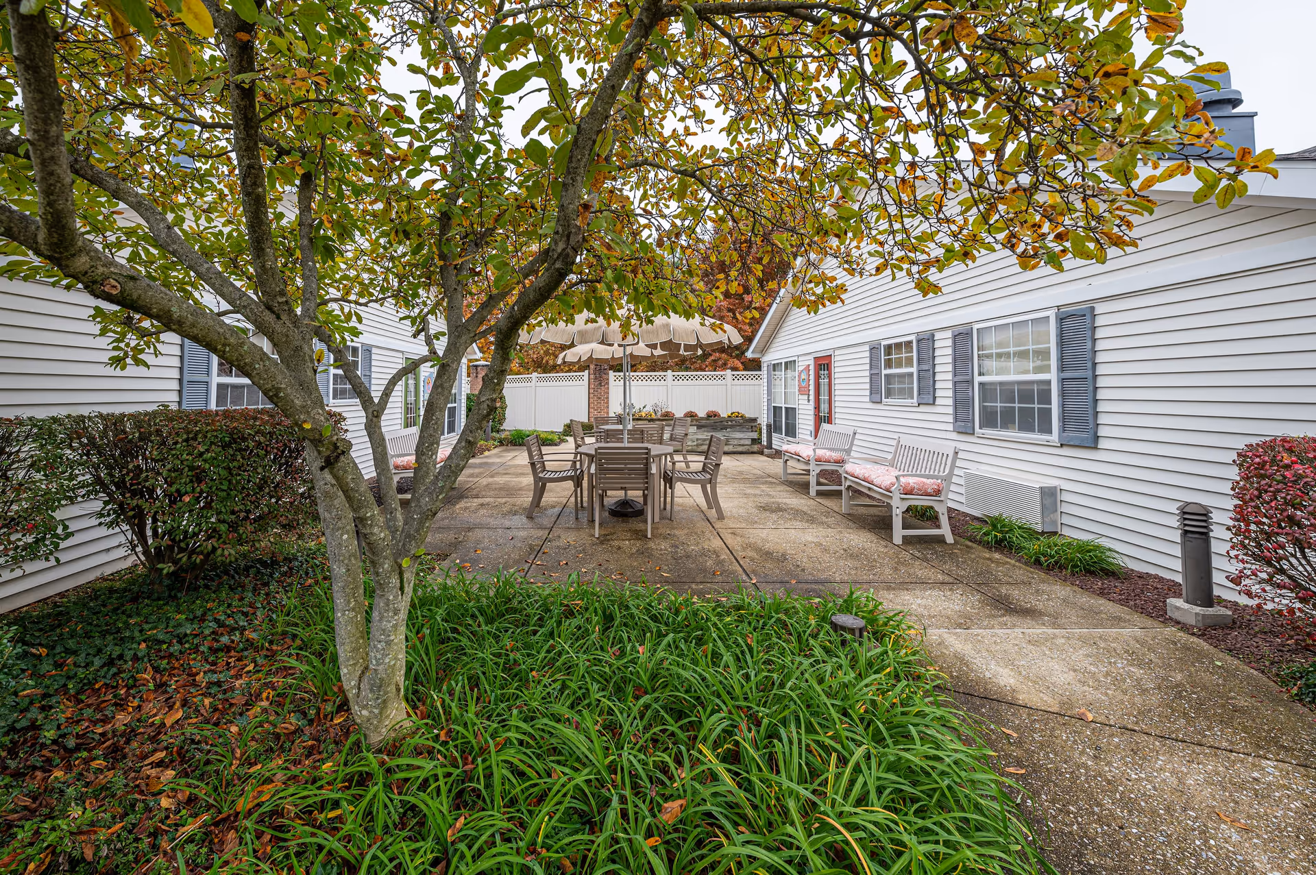 Outdoor patio area between two white buildings with blue shutters. The patio has a table with chairs and an umbrella, along with benches with cushions. There are trees and bushes surrounding the patio area.