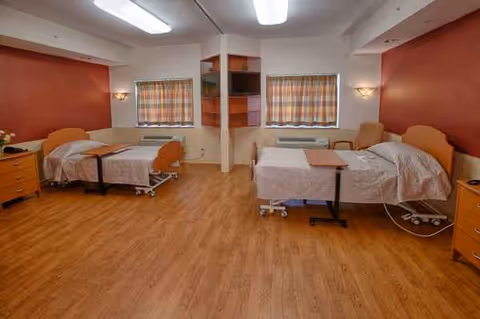Shared bedroom in a nursing facility with two beds, curtains, and polished wood flooring.