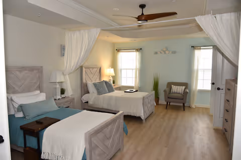 A bright and spacious bedroom with two single beds featuring light wood headboards and footboards. Each bed is made with white and teal bedding and has a small bedside table with a lamp. The room has light wood flooring, a ceiling fan, two windows with sheer curtains, a gray armchair with a cushion, and a tall dresser. White curtains hang from a ceiling track, partially enclosing the beds.