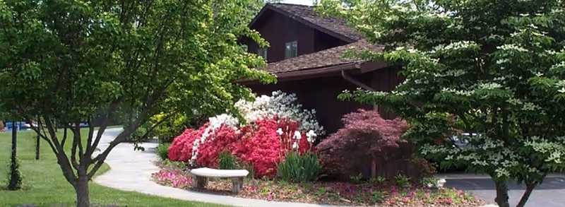 A landscaped garden area with a curved concrete pathway, colorful flowering bushes including pink and white azaleas, a stone bench, and a dark brown wooden building partially visible behind the greenery.