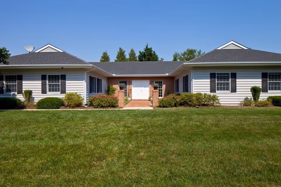Single-story building with white siding and black shutters, a central brick entrance with a white door, surrounded by green bushes and a well-maintained lawn under a clear blue sky.