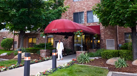 Entrance of a brick building with a red canopy displaying the number 27577. A person dressed in white is walking on the sidewalk leading from the entrance. The area is landscaped with green grass, flowers, bushes, and trees.