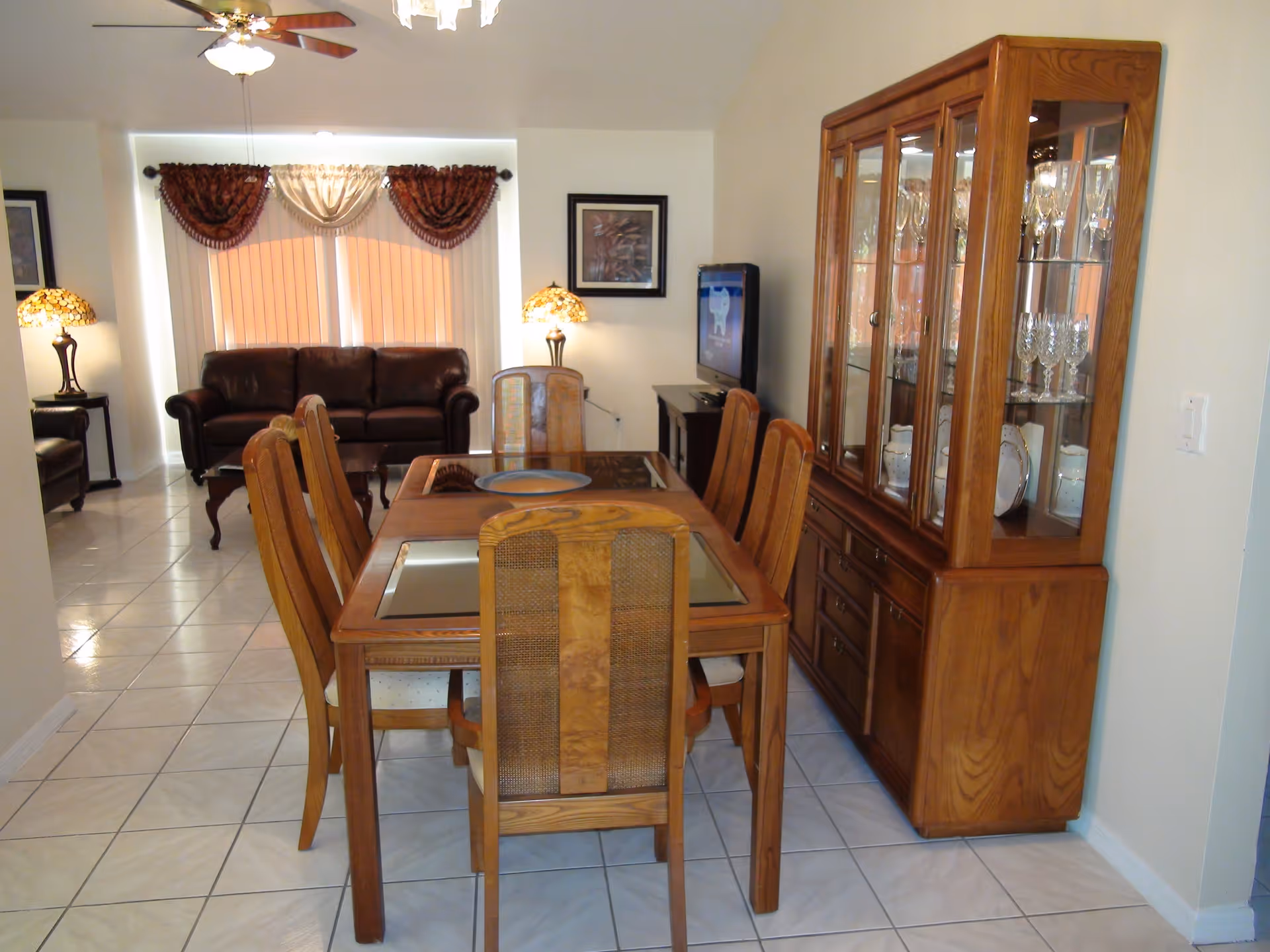 A dining room with a wooden dining table and six chairs. To the right, there is a wooden china cabinet displaying glassware and plates. In the background, there is a living area with a dark brown leather sofa, two side tables with lamps, a ceiling fan, and a TV on a stand. The floor is tiled with white tiles, and the windows have vertical blinds with decorative valances.