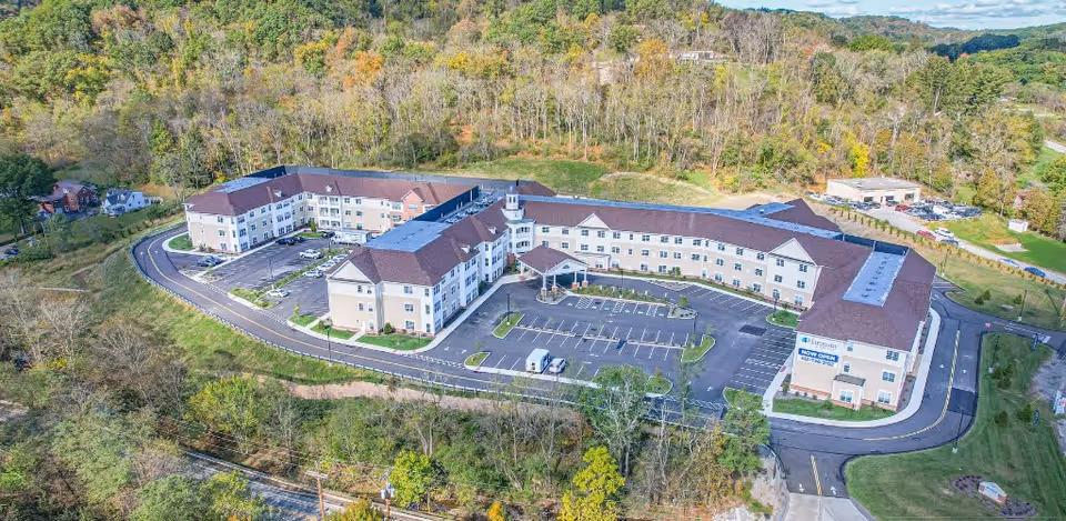 Aerial view of a large U-shaped senior living building with a central entrance, parking lot, and surrounding wooded landscape.