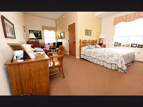 A cozy bedroom in a senior living facility with a wooden bed covered in a floral bedspread, a wooden desk and chair, a red armchair, a small TV on a stand, and framed pictures on the walls and windowsill. The room has beige walls and carpeted floor.