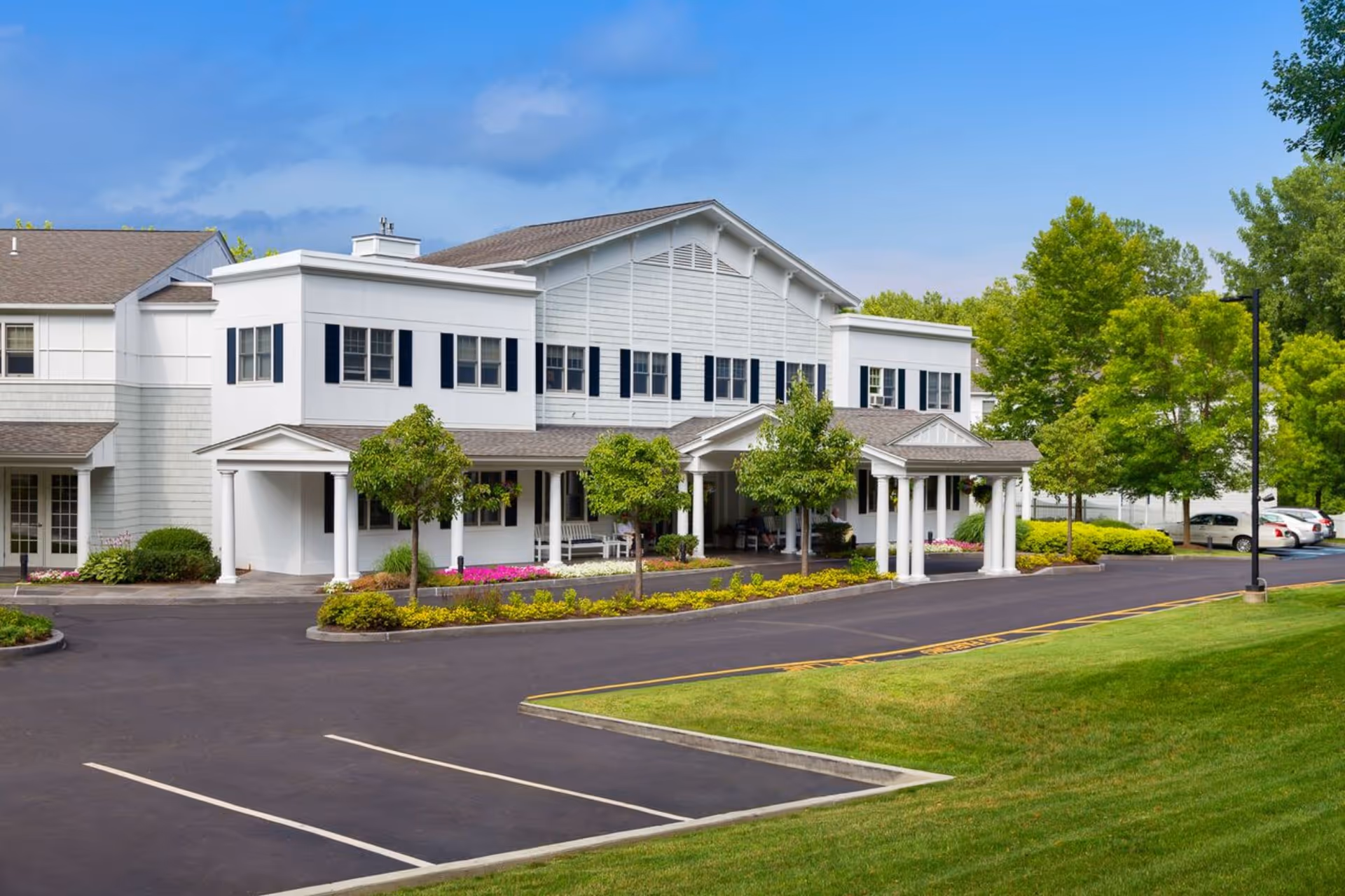 Exterior view of a two-story senior living facility building with white siding and black shutters, surrounded by green trees and landscaping, with a paved driveway and parking area in front under a partly cloudy blue sky.