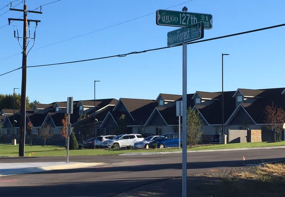 Street view of a senior living complex showing multiple connected peaked-roof buildings, parked cars, and a street sign at the corner under a clear blue sky.