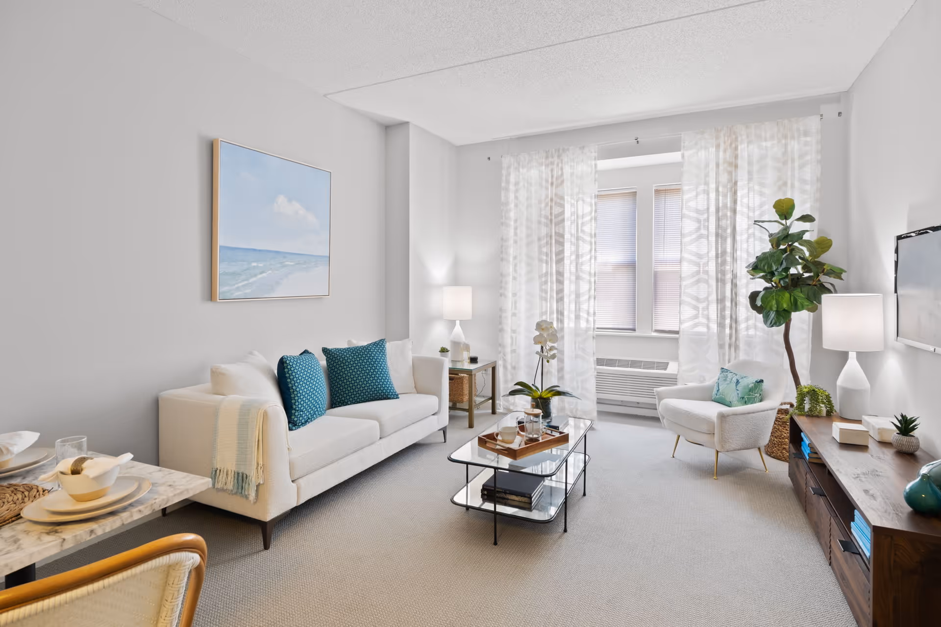 A bright and modern living room with a white sofa adorned with teal cushions, a glass coffee table with decorative items, a white armchair with a cushion, a wooden TV stand with a mounted flat-screen TV, two table lamps, a large potted plant, and sheer patterned curtains covering a window.
