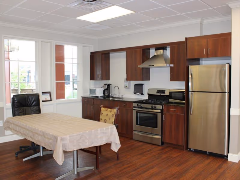 Community kitchenette and dining area with stainless steel appliances, dark wood cabinets, a table with a tablecloth and chairs, and large windows.