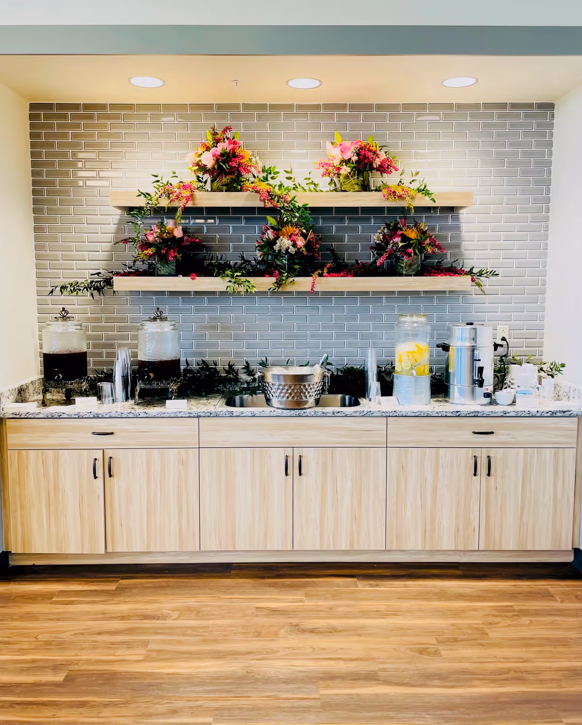 A beverage station with a granite countertop and light wood cabinets below. On the counter are two large glass dispensers with dark liquid, a dispenser with lemon water, a metal ice bucket, a coffee urn, and several clear plastic cups. Above the counter are two wooden shelves mounted on a gray tiled wall, decorated with vibrant floral arrangements and greenery. The floor is wood with a natural finish.
