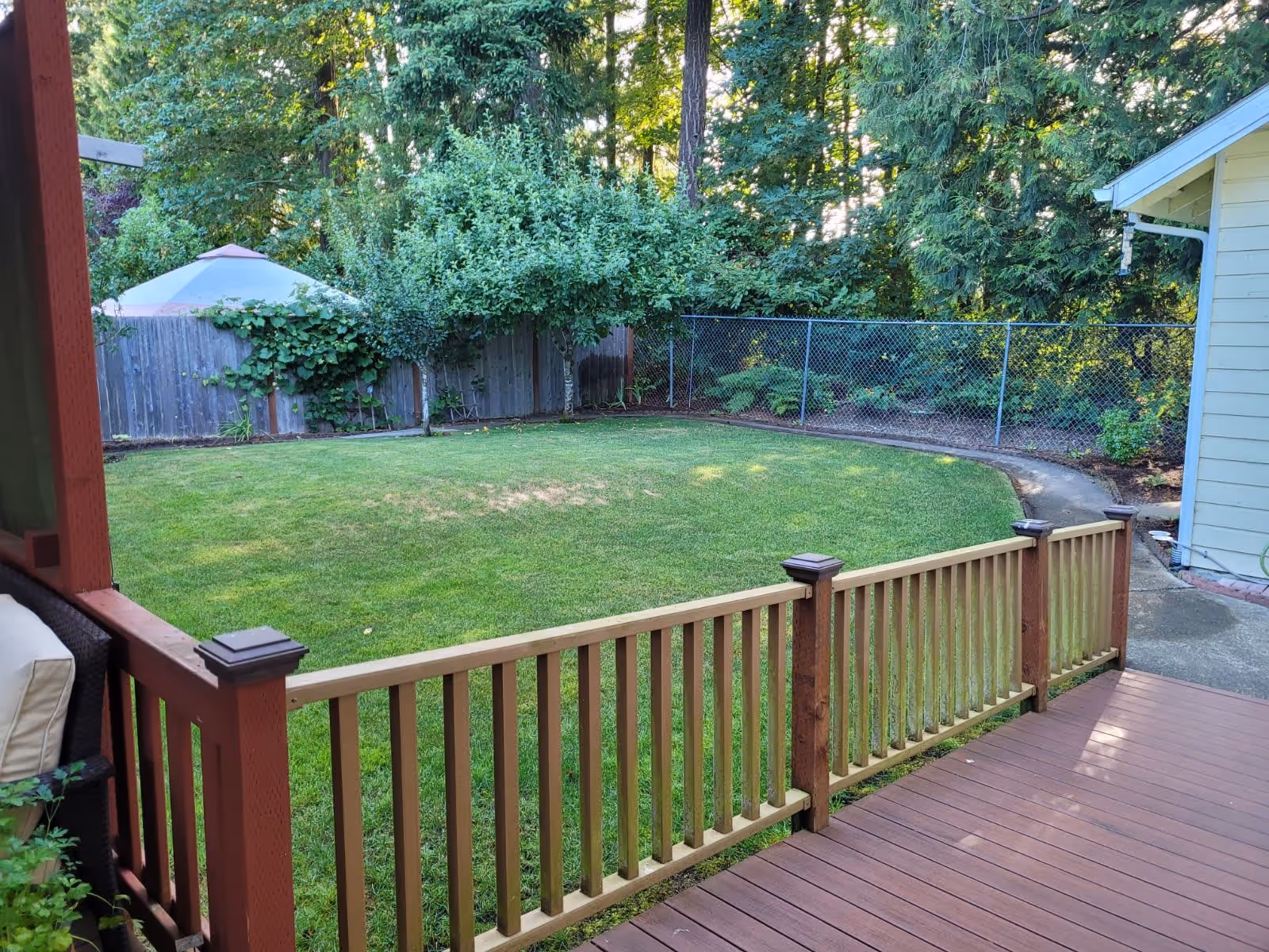 View of a backyard garden area with a wooden deck and railing in the foreground, a well-maintained grassy lawn, small trees, and a wooden fence with a chain-link fence behind it. The area is surrounded by tall trees and greenery.