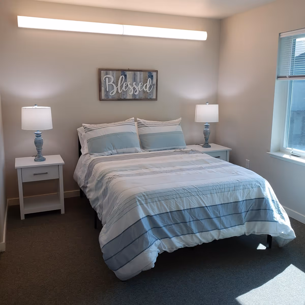 A neatly made bed with striped bedding between two nightstands and lamps under a wall sign reading 'Blessed' in a bright bedroom.