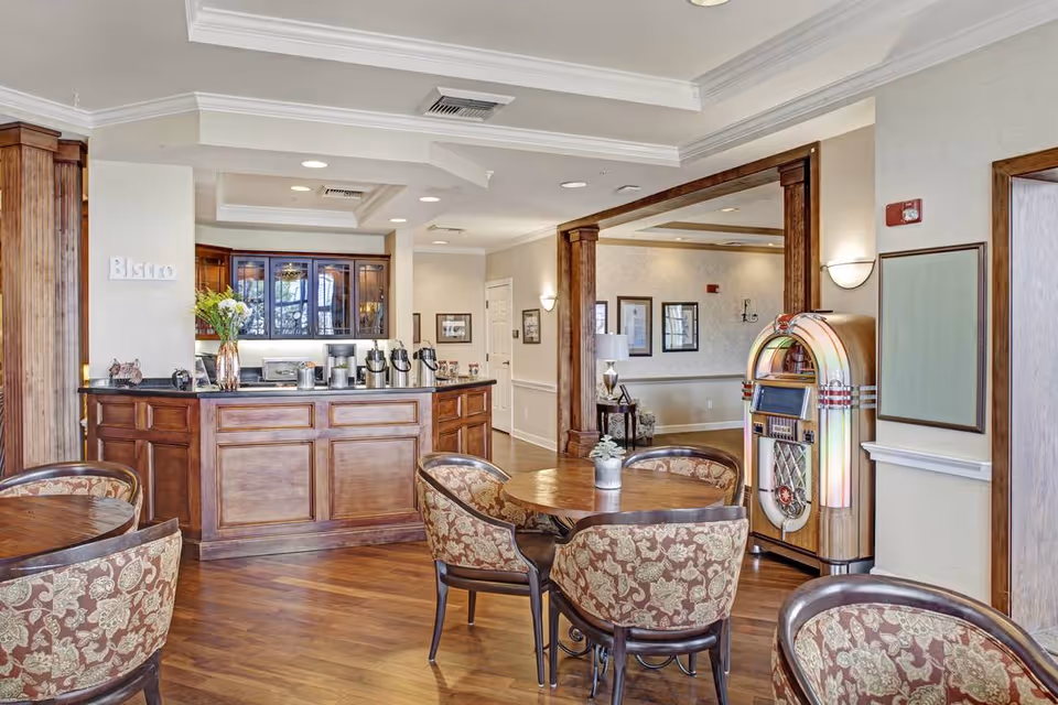 Cozy bistro-style dining area with a wooden service counter, round tables and upholstered chairs, and a jukebox against the wall.