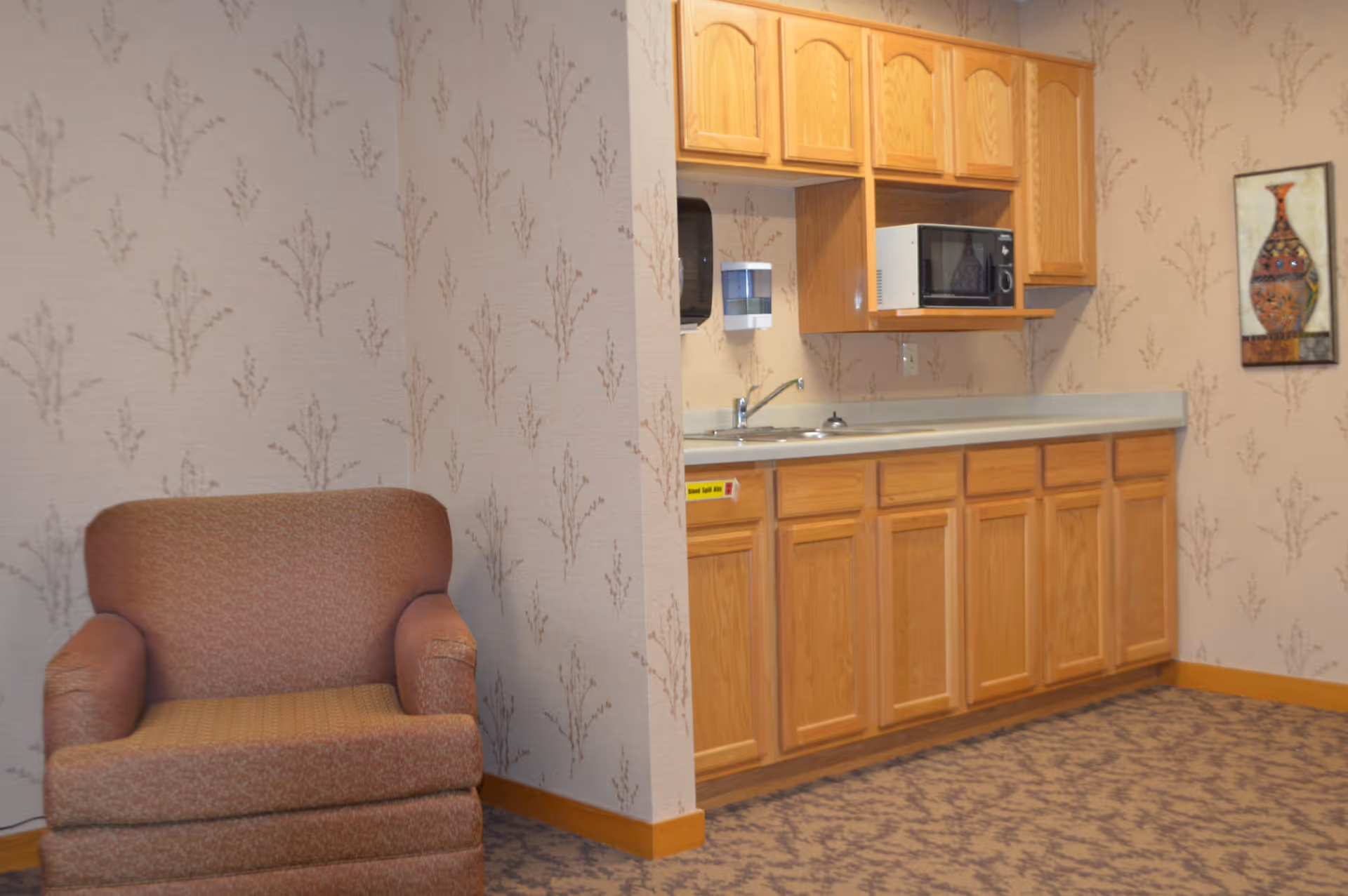 Small kitchenette with wooden cabinets, a sink and microwave next to an upholstered armchair in a carpeted room.
