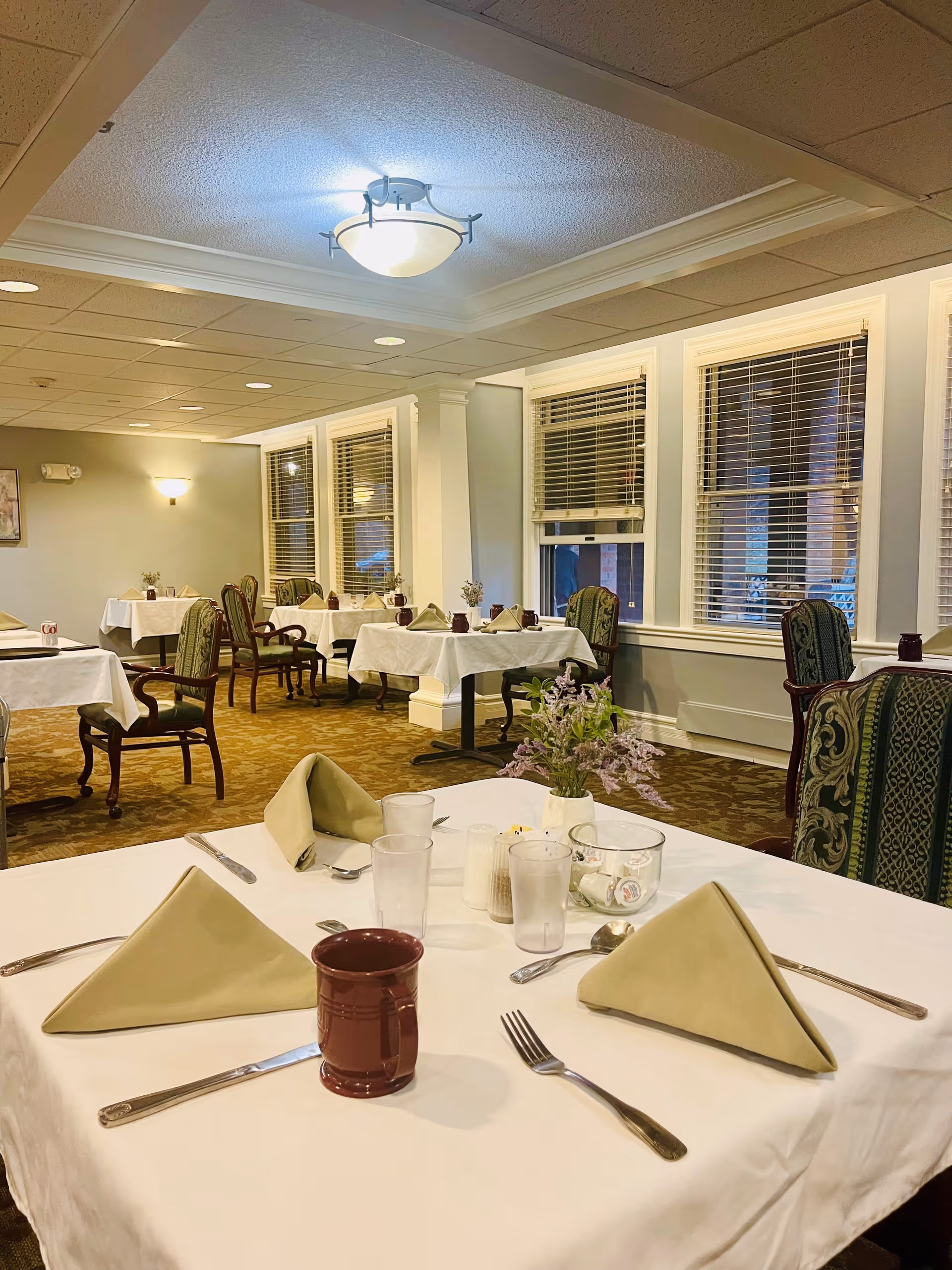 Dining room with tables covered in white tablecloths, set with beige folded napkins, silverware, glasses, and maroon mugs. The room has patterned carpet, green upholstered chairs, large windows with blinds, and soft ceiling lighting.