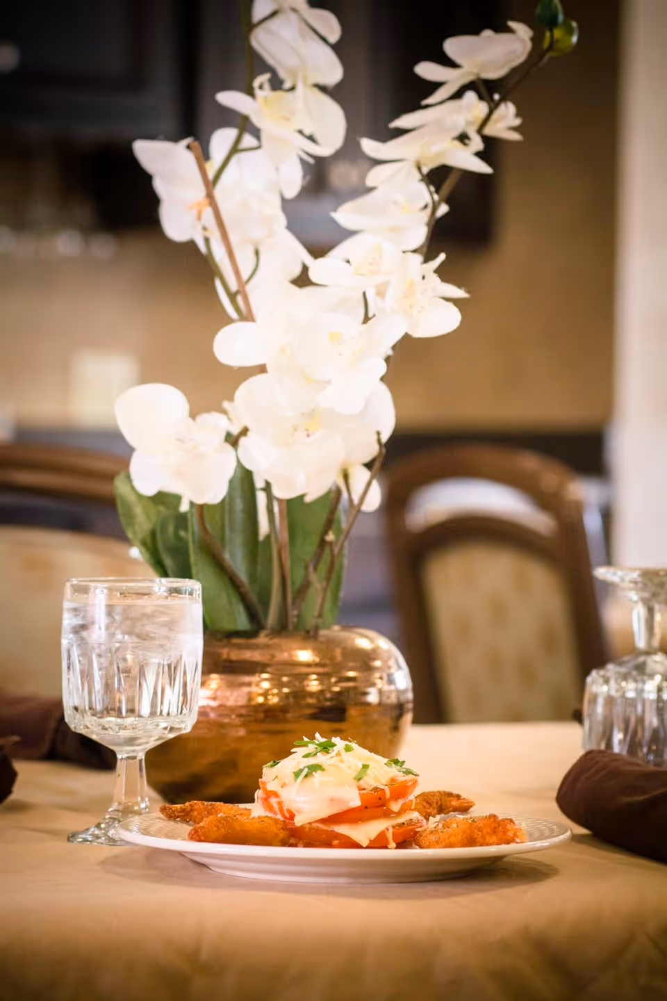A dining table set with a plate of food featuring fried items topped with sliced tomatoes and melted cheese garnished with herbs. Next to the plate is a glass of water with ice. In the background, there is a decorative vase with white orchid flowers and blurred dining chairs.