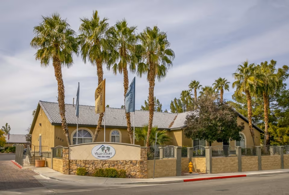 Front exterior of Villa Court Assisted Living, a single-story building with palm trees, flags, and a stone entrance sign.