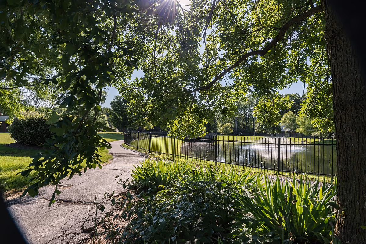 A sunny outdoor scene at Altenheim Family-first Senior Living featuring a paved walking path bordered by green bushes and trees. A black metal fence runs alongside a pond with a water fountain in the background. Sunlight filters through the tree leaves, creating a peaceful and inviting atmosphere.
