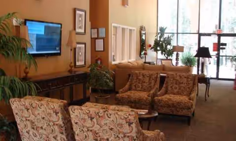 Spacious senior living lounge with patterned chairs, plants, and a wall-mounted TV.
