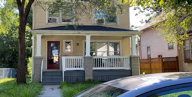 Front exterior view of a two-story beige house with a covered porch, white railing, and a red front door. There is a large tree to the left and a car parked in front of the house. The house is situated between two other houses with a wooden fence on the right side.