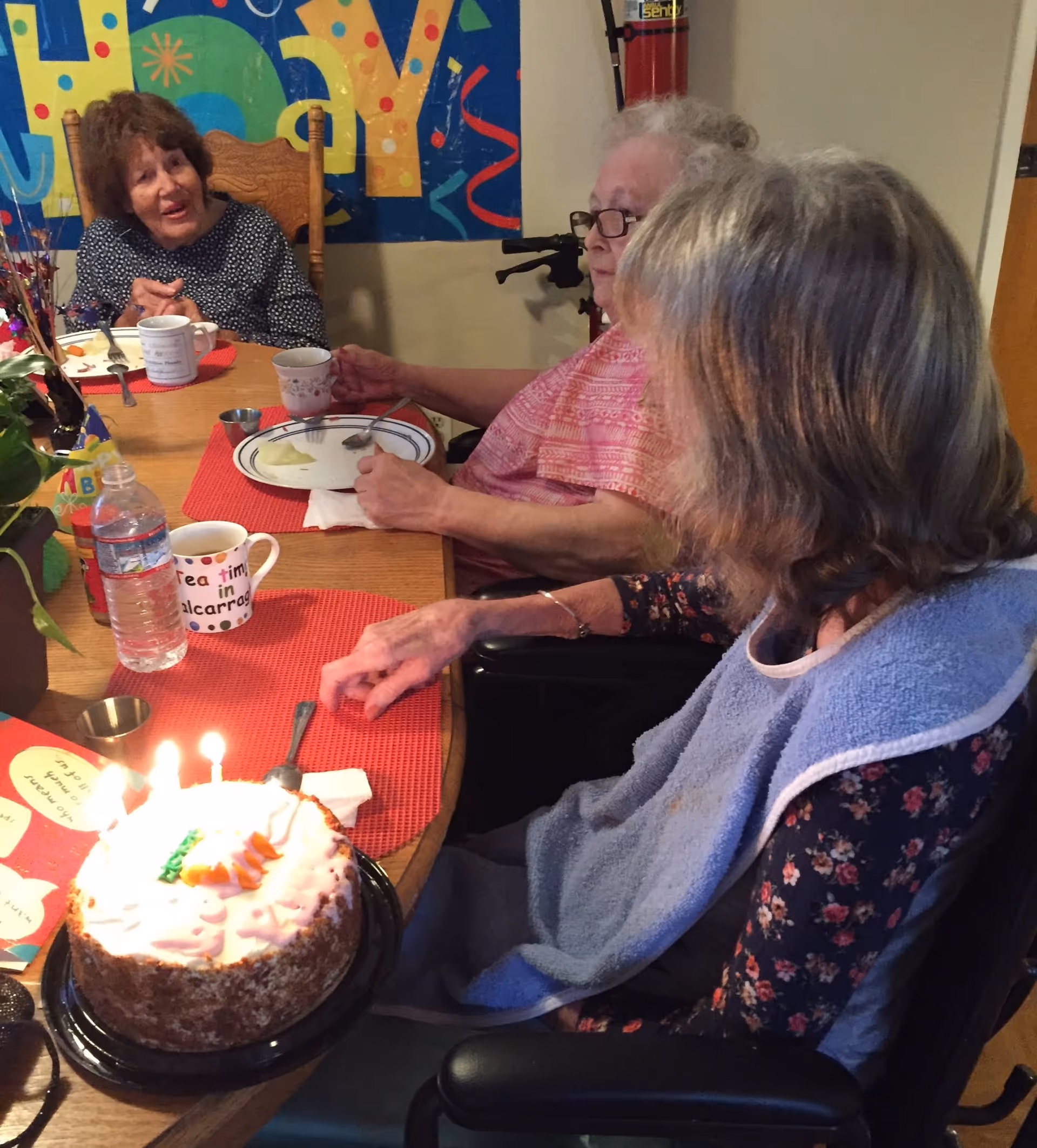 Three elderly women sitting around a wooden dining table celebrating a birthday with a cake that has lit candles. One woman is wearing a blue bib, and there is a colorful 'Happy Birthday' banner on the wall behind them. The table has red placemats, mugs, a water bottle, and some plates with food remnants.