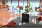 Dining room with set tables, folded napkins, glassware, and a floral centerpiece near large windows.