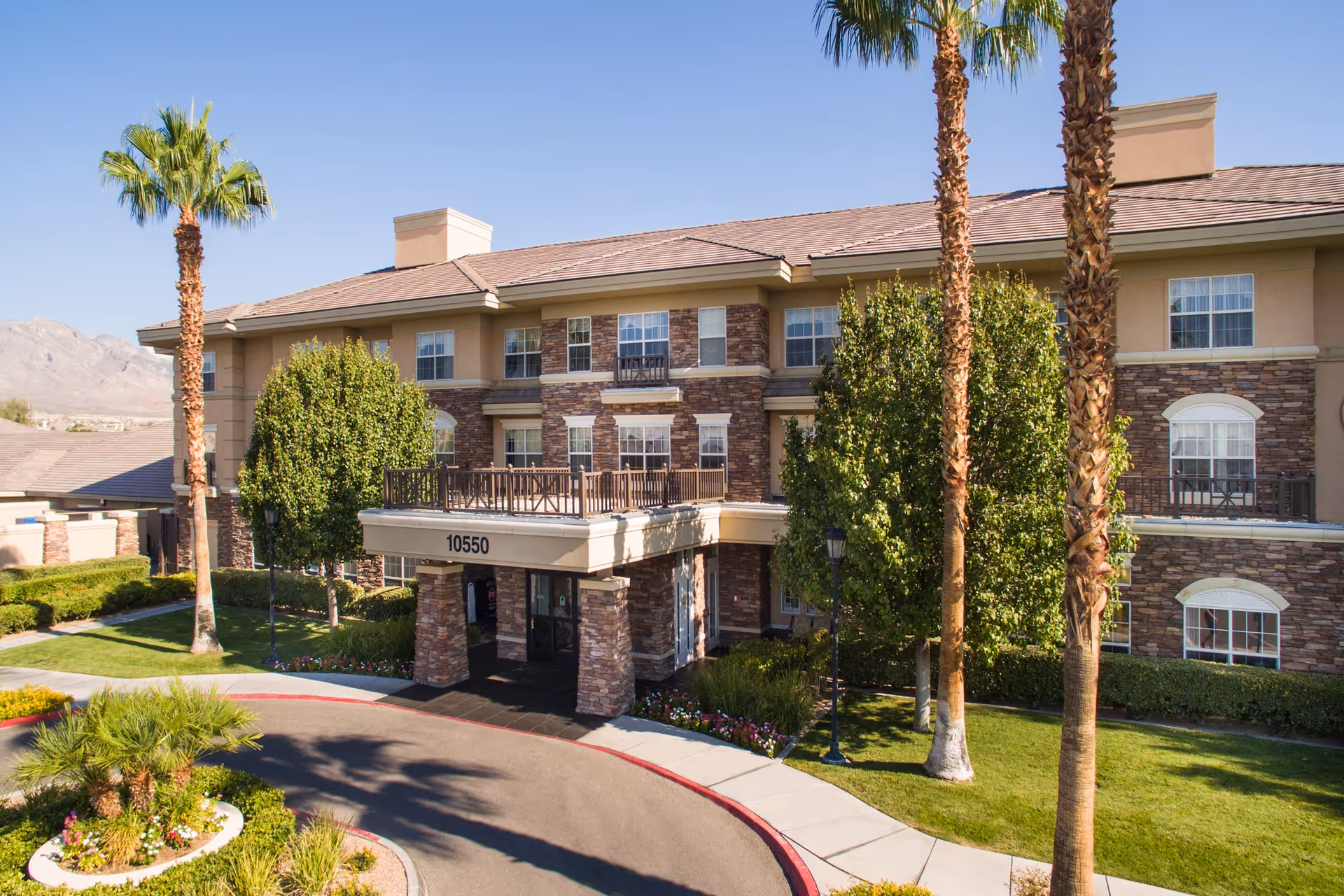 Exterior view of a three-story senior living facility building with stone and beige siding, surrounded by palm trees and well-maintained landscaping under a clear blue sky. The entrance has a covered drop-off area with the number 10550 displayed above it.